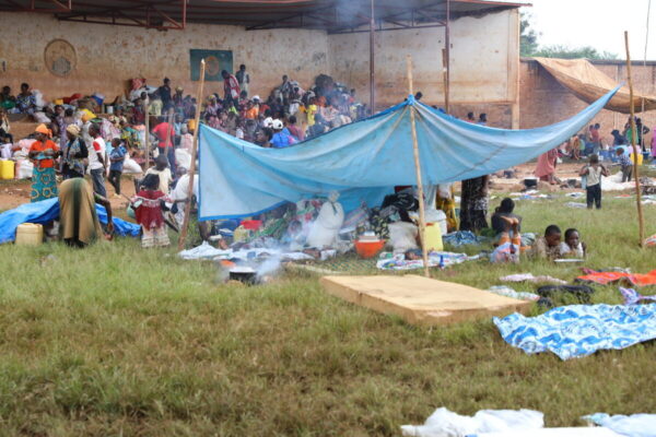 Dozens of people are seen camped out in a field near a building with  one family in the foreground sheltering with their belongings under an improvised tent made of a large light-blue tarp attached on the four corners to sticks driven into the grassy ground.