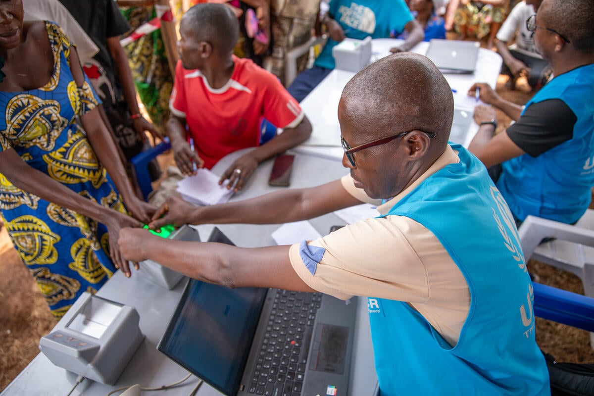 Un homme noir, chauve et d'âge moyen, portant des lunettes et un gilet de visibilité bleu clair du HCR, aide une femme portant une robe colorée à faire scanner ses empreintes digitales dans un centre d'enregistrement très fréquenté mis en place par le HCR.